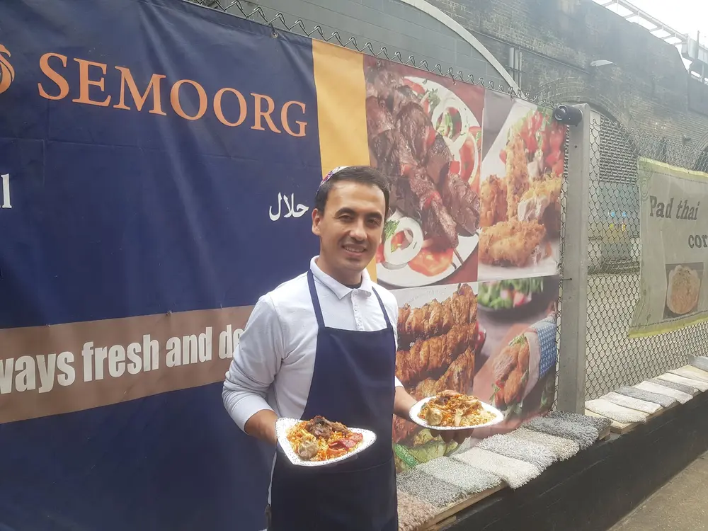Man in a Semoorg apron smiling with two plates of food in front of the stall's banner